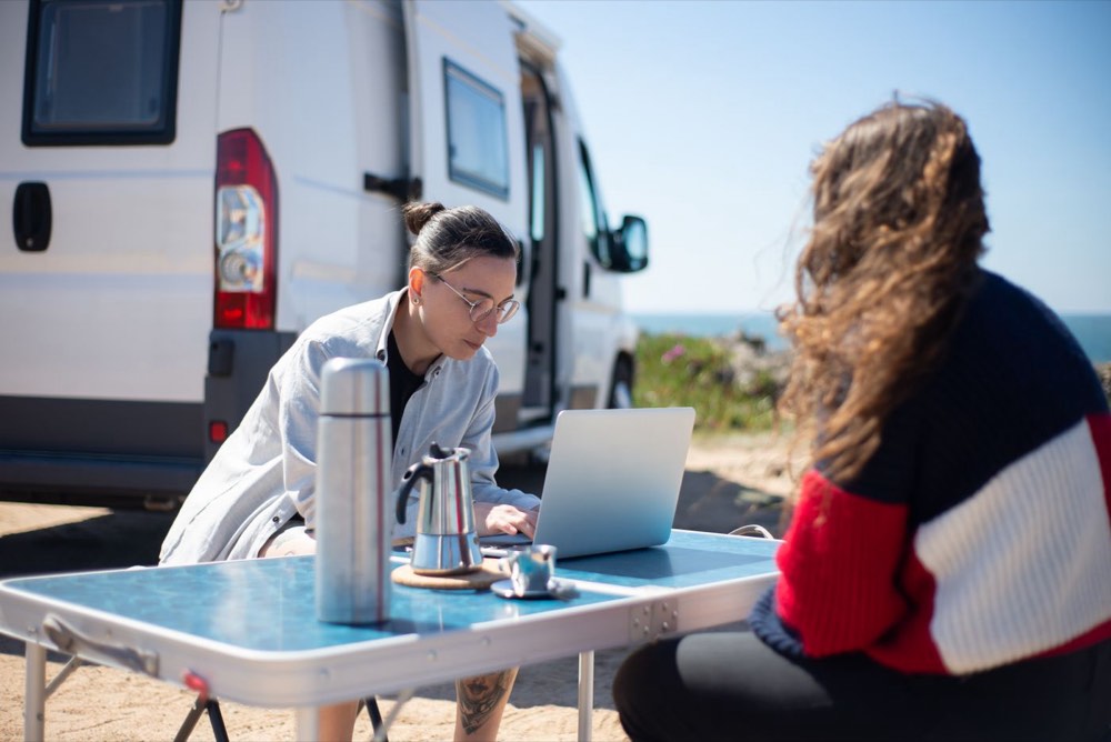 Two people working on a laptop at a folding table outside their camper van