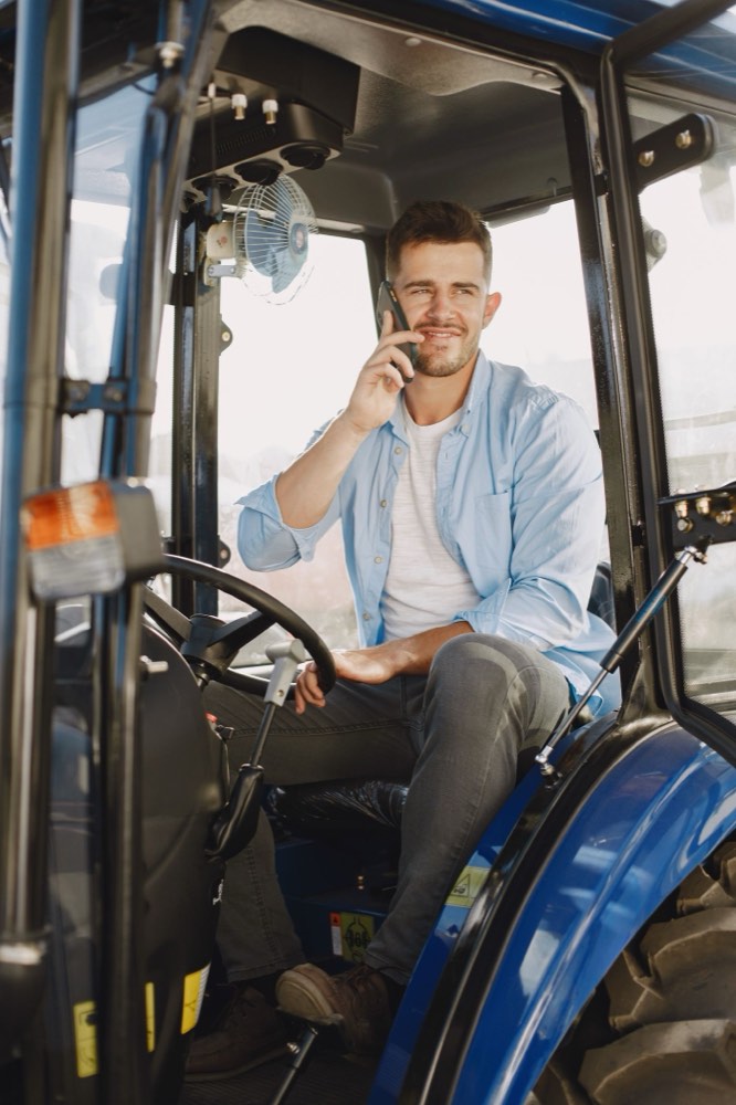 A man in a work shirt taking a phone call from the cab of his tractor