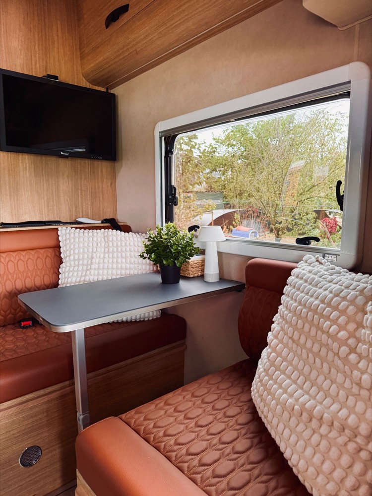 A warm, comfortable RV living area with a table, cushions and a window onto the trees