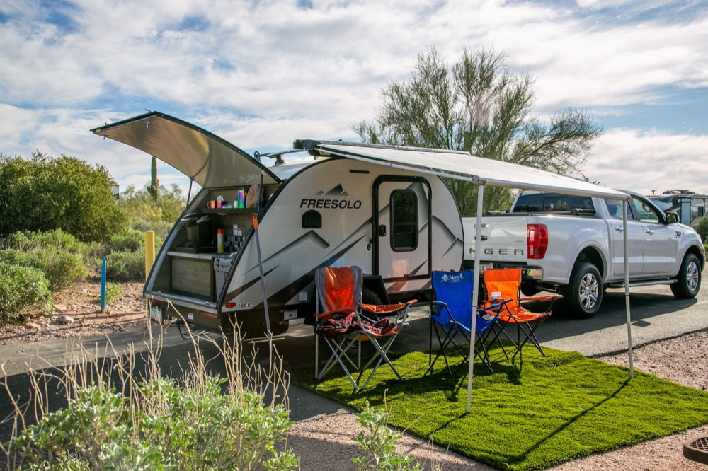 A teardrop trailer and pickup truck settled on a site with chairs and a patio mat